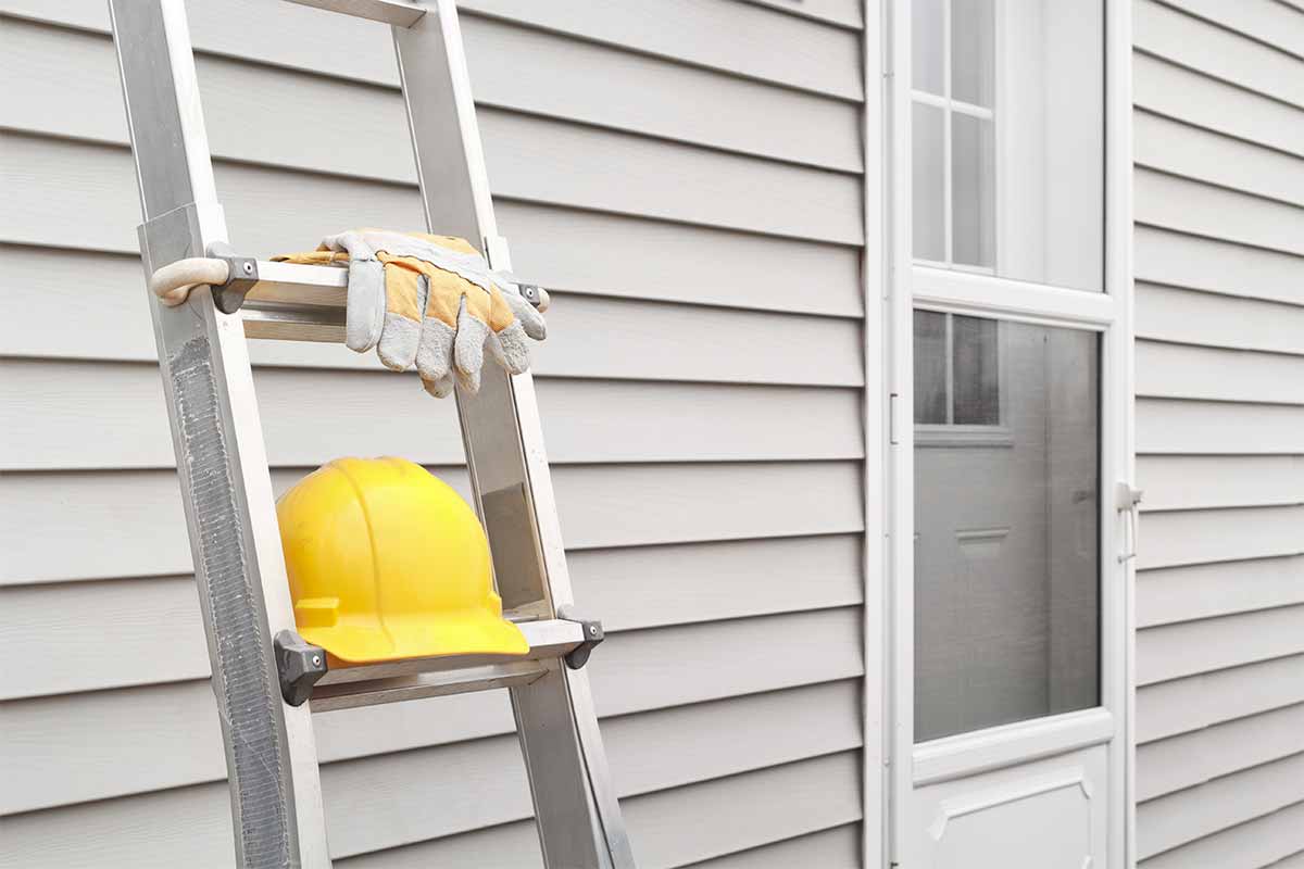 Metal ladder with work gloves and yellow hard hat leaning against white vinyl siding